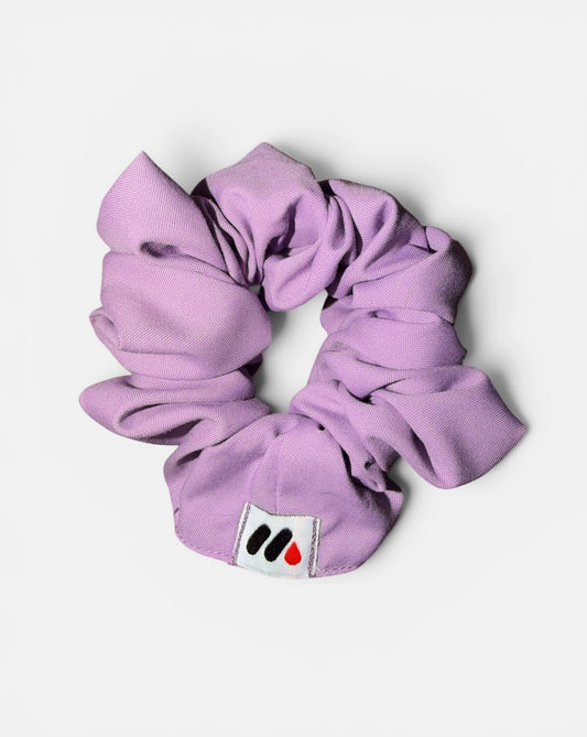 A lavender fabric hair scrunchie with a small branded white tag featuring a black and red abstract mark against a white background.
