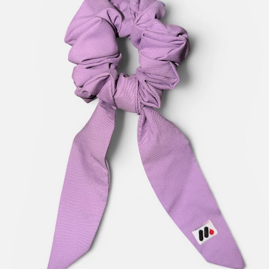 A lavender fabric hair scrunchie with long tails and a small branded white tag featuring a black and red logo against a white background.