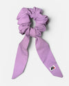 A lavender fabric hair scrunchie with long tails and a small branded white tag featuring a black and red logo against a white background.