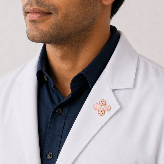 A close-up of a male healthcare professional wearing a white lab coat over a dark blue collared shirt with a pink adhesive bandage enamel lapel pin against a plain background.