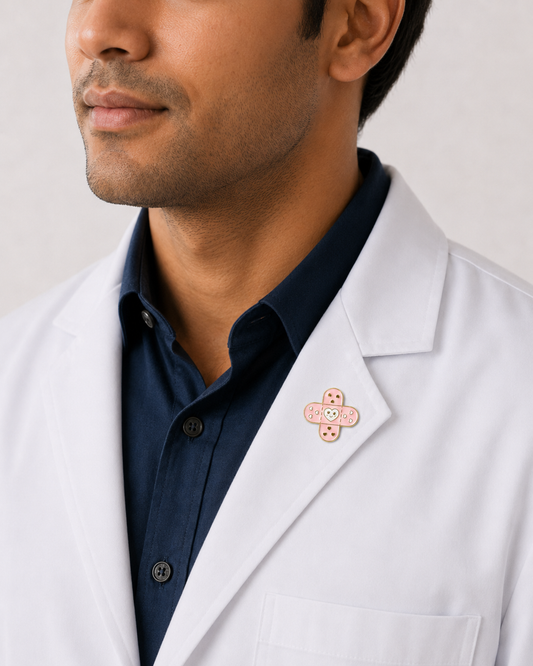 A close-up of a male healthcare professional wearing a white lab coat over a dark blue collared shirt with a pink adhesive bandage enamel lapel pin against a plain background.