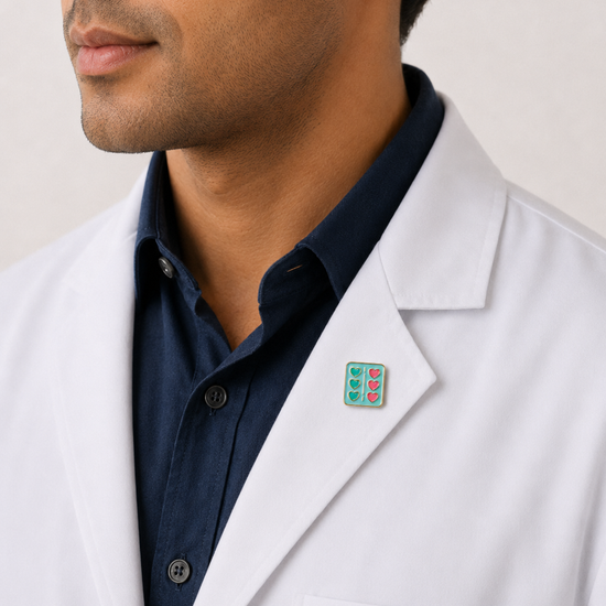 A close-up of a male healthcare professional wearing a white lab coat over a dark blue collared shirt with a decorative enamel lapel pin against a plain background.