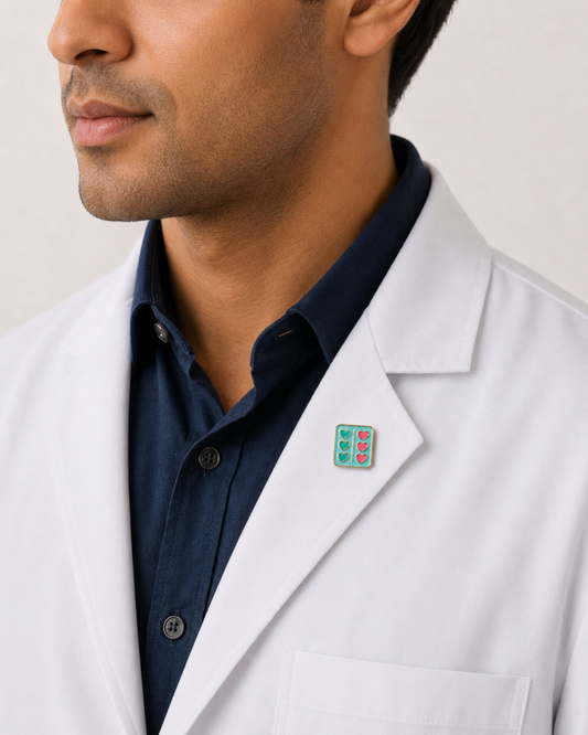 A close-up of a male healthcare professional wearing a white lab coat over a dark blue collared shirt with a decorative enamel lapel pin against a plain background.