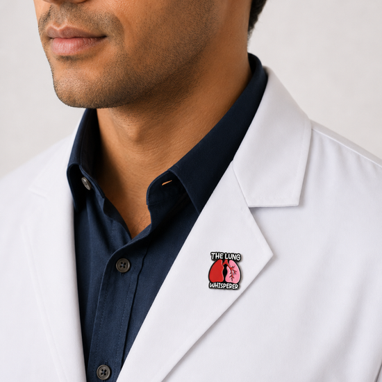 A close-up of a male healthcare professional wearing a white lab coat over a dark blue collared shirt with a "The Lung Whisperer" enamel lapel pin against a plain background.