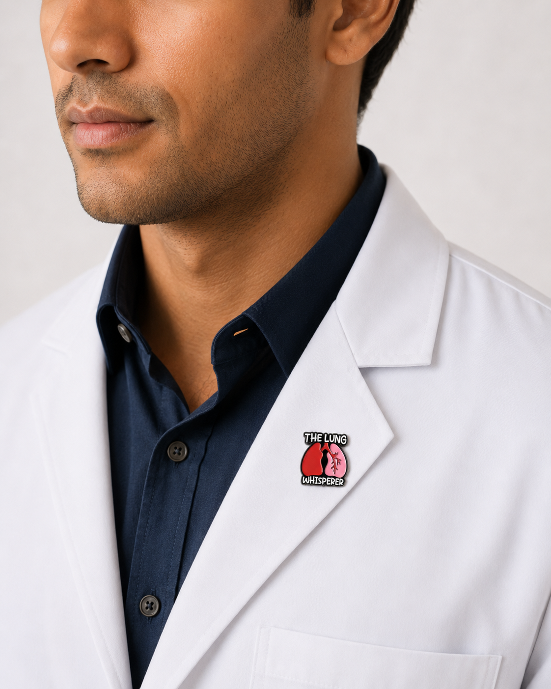 A close-up of a male healthcare professional wearing a white lab coat over a dark blue collared shirt with a "The Lung Whisperer" enamel lapel pin against a plain background.