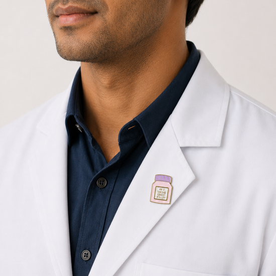 A close-up of a male healthcare professional wearing a white lab coat over a dark blue collared shirt with a purple and white medicine pill bottle enamel lapel pin against a plain background.