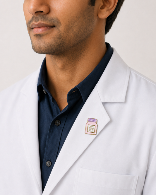 A close-up of a male healthcare professional wearing a white lab coat over a dark blue collared shirt with a purple and white medicine pill bottle enamel lapel pin against a plain background.