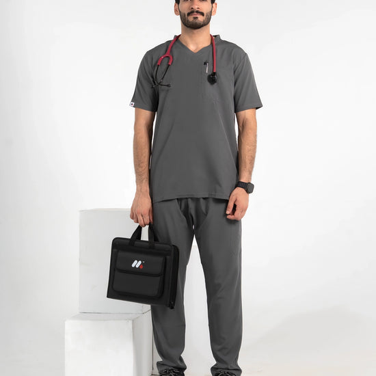 A male model wearing a dark gray medical scrub set with a V-neck top and matching pants, holding a black medical bag and wearing a stethoscope, MedFit.