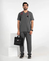 A male model wearing a dark gray medical scrub set with a V-neck top and matching pants, holding a black medical bag and wearing a stethoscope, MedFit.