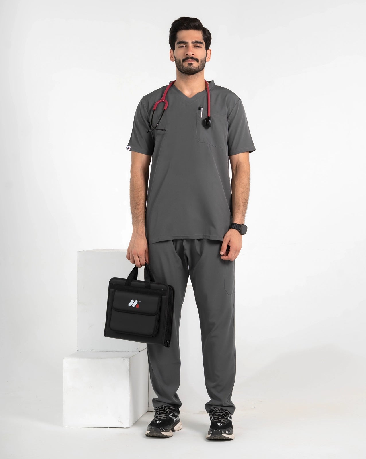 A male model wearing a dark gray medical scrub set with a V-neck top and matching pants, holding a black medical bag and wearing a stethoscope, MedFit.