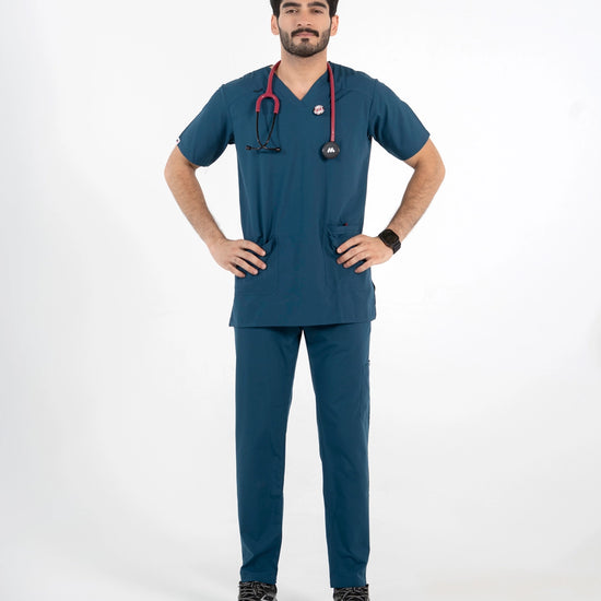 A male model posing in a two-piece dark teal-blue medical scrub set featuring a short-sleeve V-neck top and matching straight-leg pants, wearing a red stethoscope, MedFit.