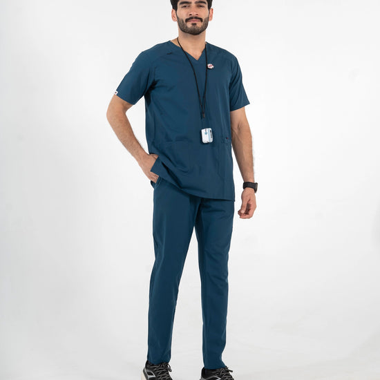 A male model wearing a dark teal blue V-neck scrub set with matching pants, paired with black sneakers and a black lanyard with an ID badge, MedFit professional workwear.