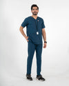 A male model wearing a dark teal blue V-neck scrub set with matching pants, paired with black sneakers and a black lanyard with an ID badge, MedFit professional workwear.