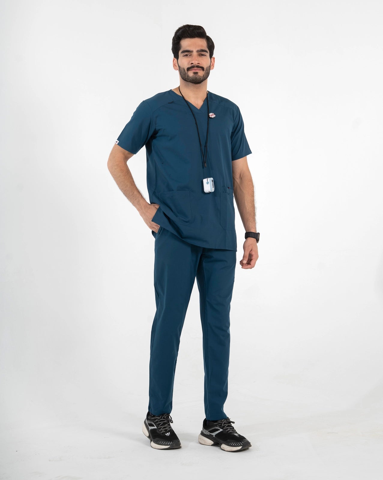 A male model wearing a dark teal blue V-neck scrub set with matching pants, paired with black sneakers and a black lanyard with an ID badge, MedFit professional workwear.