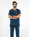 A male model with arms crossed wearing a dark teal-blue V-neck scrub top and matching pants, equipped with a red stethoscope, MedFit professional medical uniform.