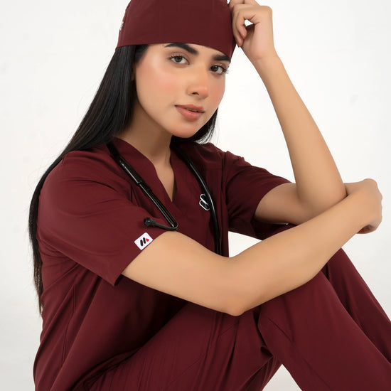 A healthcare professional sitting down in a close-up pose while wearing a maroon medical scrub set, a matching scrub cap, and a stethoscope against a white background.