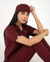 A healthcare professional sitting down in a close-up pose while wearing a maroon medical scrub set, a matching scrub cap, and a stethoscope against a white background.
