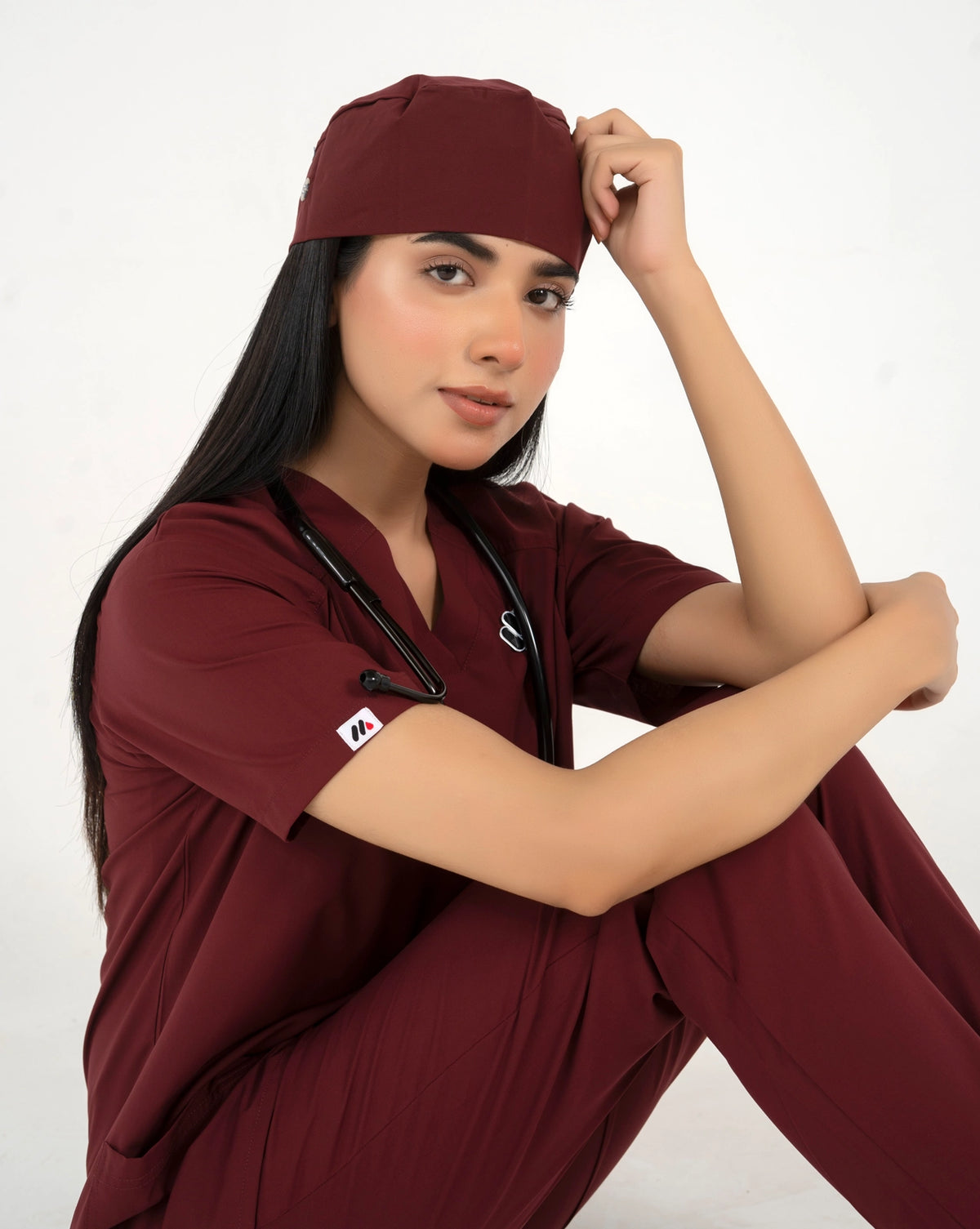 A healthcare professional sitting down in a close-up pose while wearing a maroon medical scrub set, a matching scrub cap, and a stethoscope against a white background.