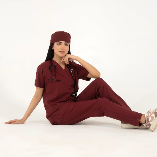 A healthcare professional sitting on the floor while modeling a maroon medical scrub set with a V-neck top, matching trousers, a matching scrub cap, and a stethoscope against a white background.