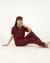 A healthcare professional sitting on the floor while modeling a maroon medical scrub set with a V-neck top, matching trousers, a matching scrub cap, and a stethoscope against a white background.