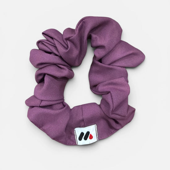 A mauve fabric hair scrunchie with a small branded white tag featuring a black and red abstract mark against a white background.