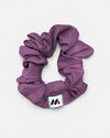 A mauve fabric hair scrunchie with a small branded white tag featuring a black and red abstract mark against a white background.