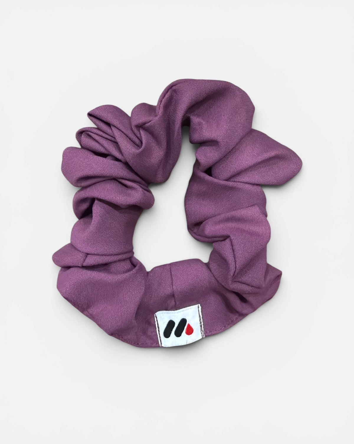 A mauve fabric hair scrunchie with a small branded white tag featuring a black and red abstract mark against a white background.