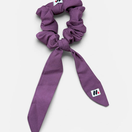 A mauve fabric hair scrunchie with long tails and a small branded white tag featuring a black and red logo against a white background.