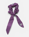A mauve fabric hair scrunchie with long tails and a small branded white tag featuring a black and red logo against a white background.