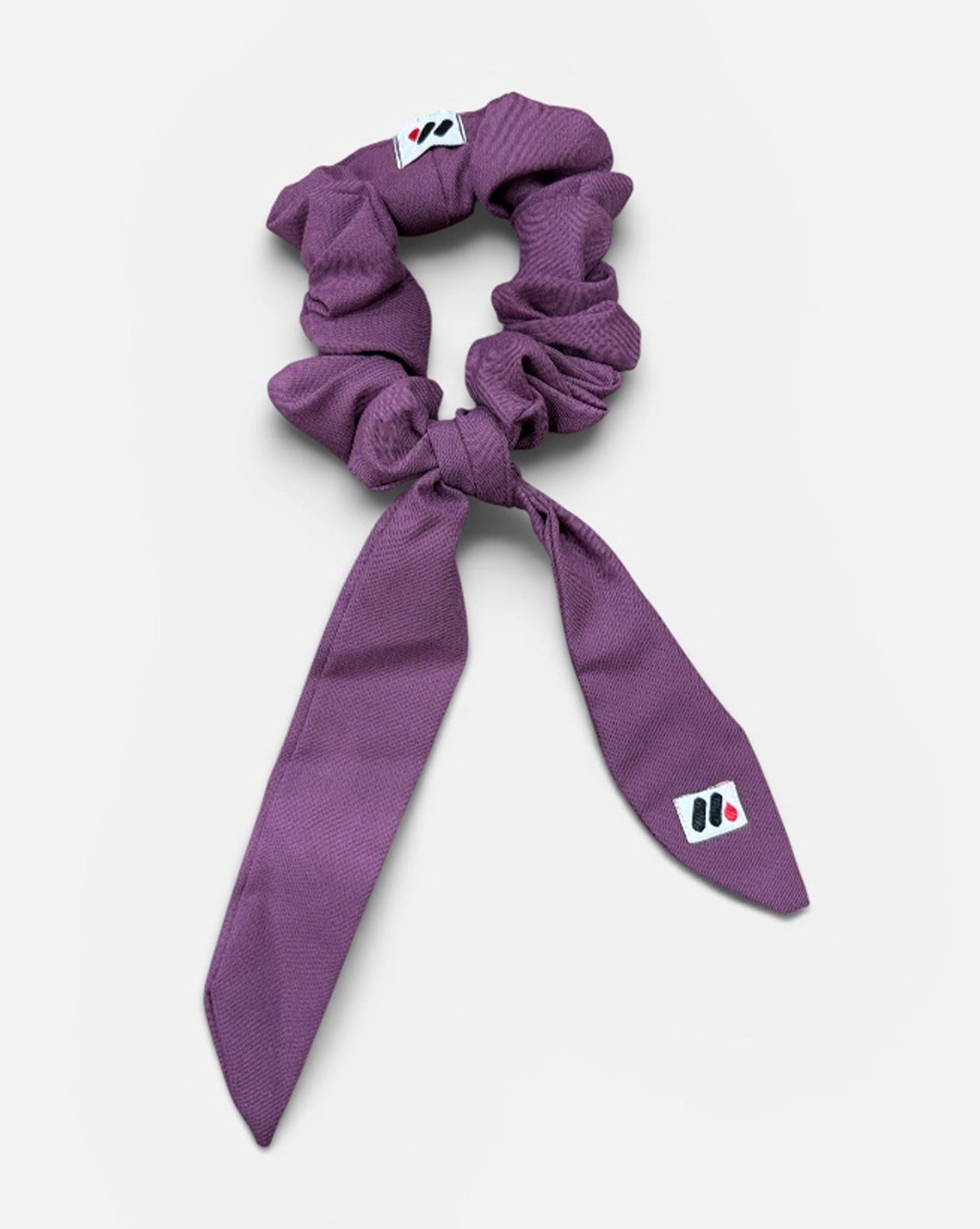 A mauve fabric hair scrunchie with long tails and a small branded white tag featuring a black and red logo against a white background.