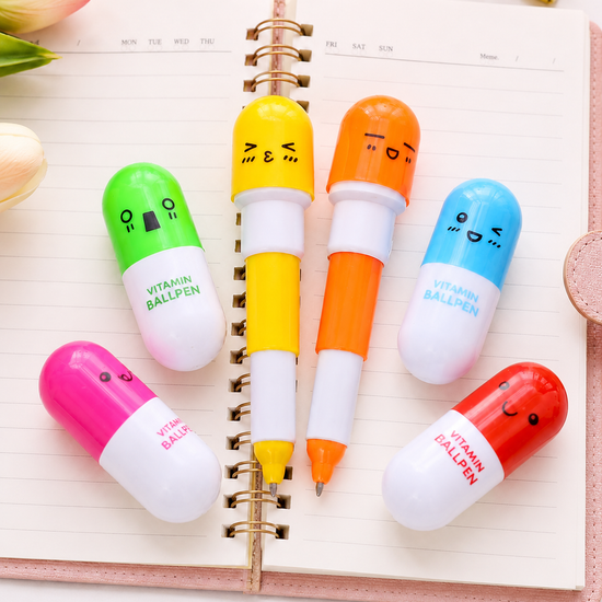 A collection of MedFit colorful pill-shaped vitamin ballpens with cute faces arranged on an open pink notebook, perfect novelty stationery for healthcare workers.