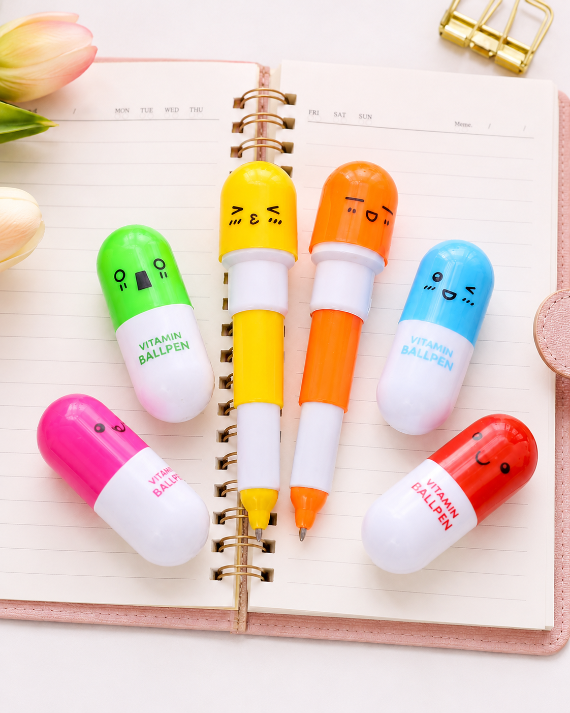 A collection of MedFit colorful pill-shaped vitamin ballpens with cute faces arranged on an open pink notebook, perfect novelty stationery for healthcare workers.