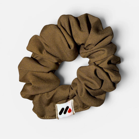 A mocha brown fabric hair scrunchie with a small branded white tag featuring black and red abstract marks against a white background.