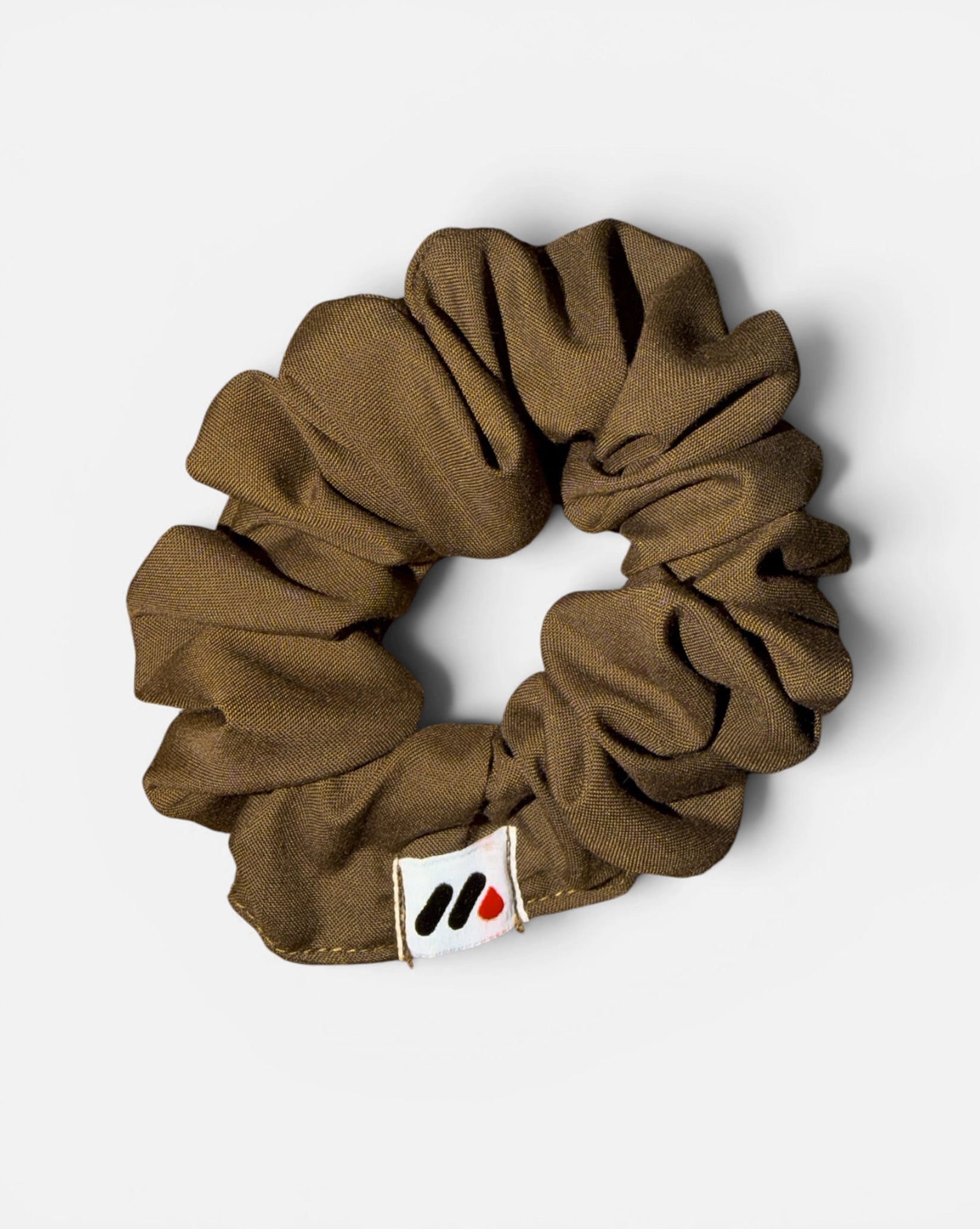 A mocha brown fabric hair scrunchie with a small branded white tag featuring black and red abstract marks against a white background.