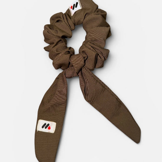 A mocha brown fabric hair scrunchie with long tails and a small branded white tag featuring a black and red logo against a white background.