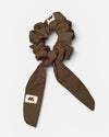A mocha brown fabric hair scrunchie with long tails and a small branded white tag featuring a black and red logo against a white background.