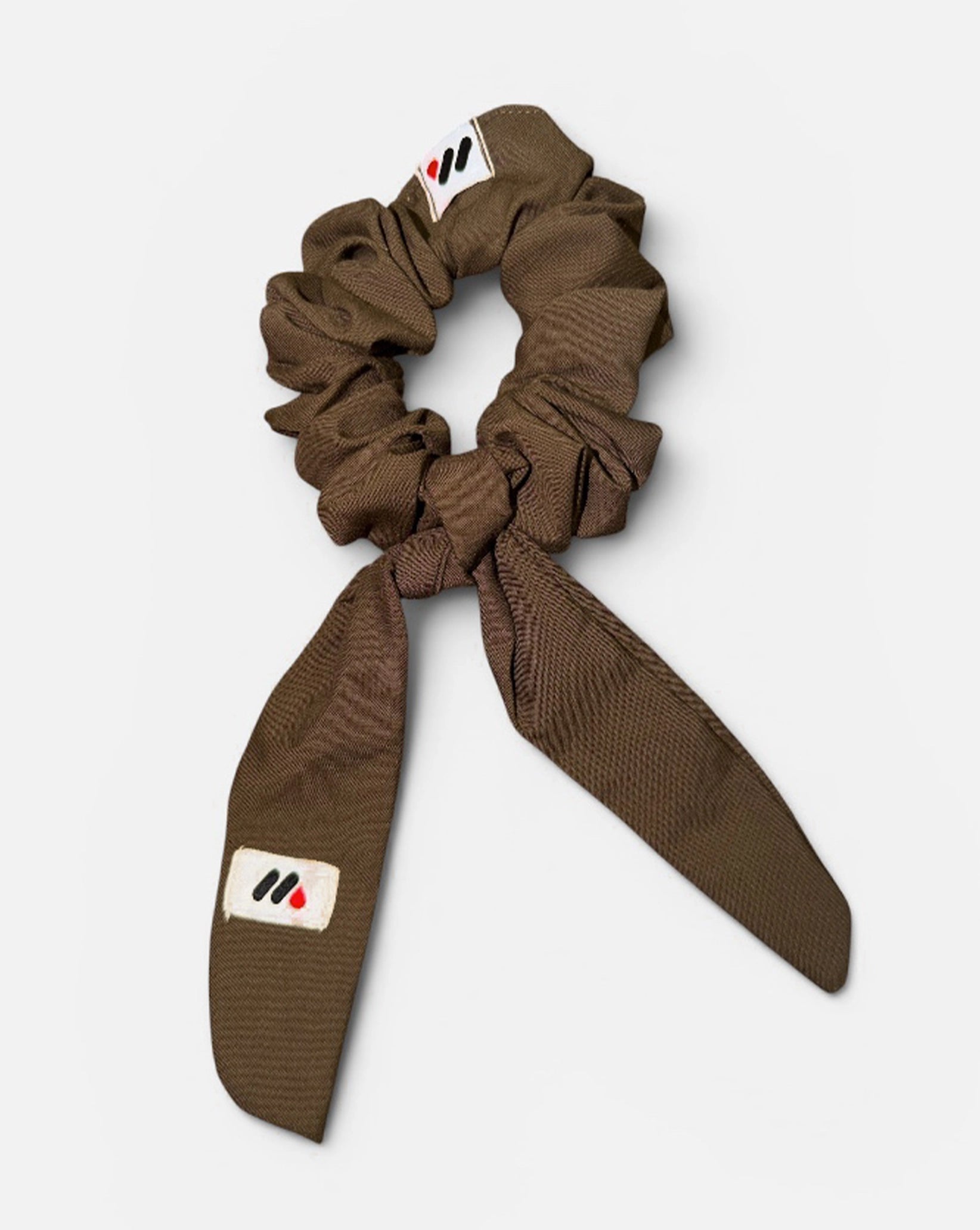 A mocha brown fabric hair scrunchie with long tails and a small branded white tag featuring a black and red logo against a white background.