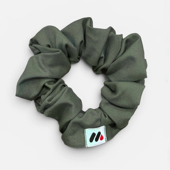 A moss green fabric hair scrunchie with a small branded white tag featuring black and red abstract marks against a white background.