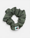 A moss green fabric hair scrunchie with a small branded white tag featuring black and red abstract marks against a white background.