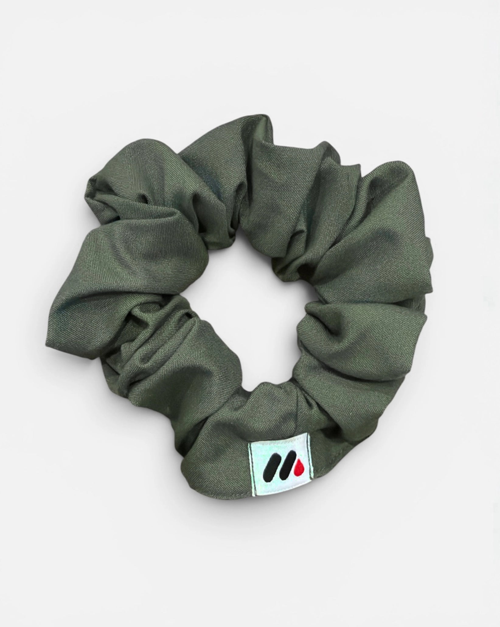 A moss green fabric hair scrunchie with a small branded white tag featuring black and red abstract marks against a white background.