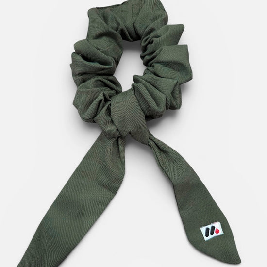 A moss green fabric hair scrunchie with long tails and a small branded white tag featuring a black and red logo against a white background.