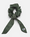 A moss green fabric hair scrunchie with long tails and a small branded white tag featuring a black and red logo against a white background.