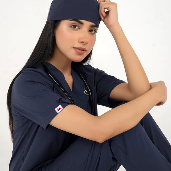 A healthcare professional wearing a matching navy blue medical scrub set with a matching scrub cap and a stethoscope, posing against a white studio background.
