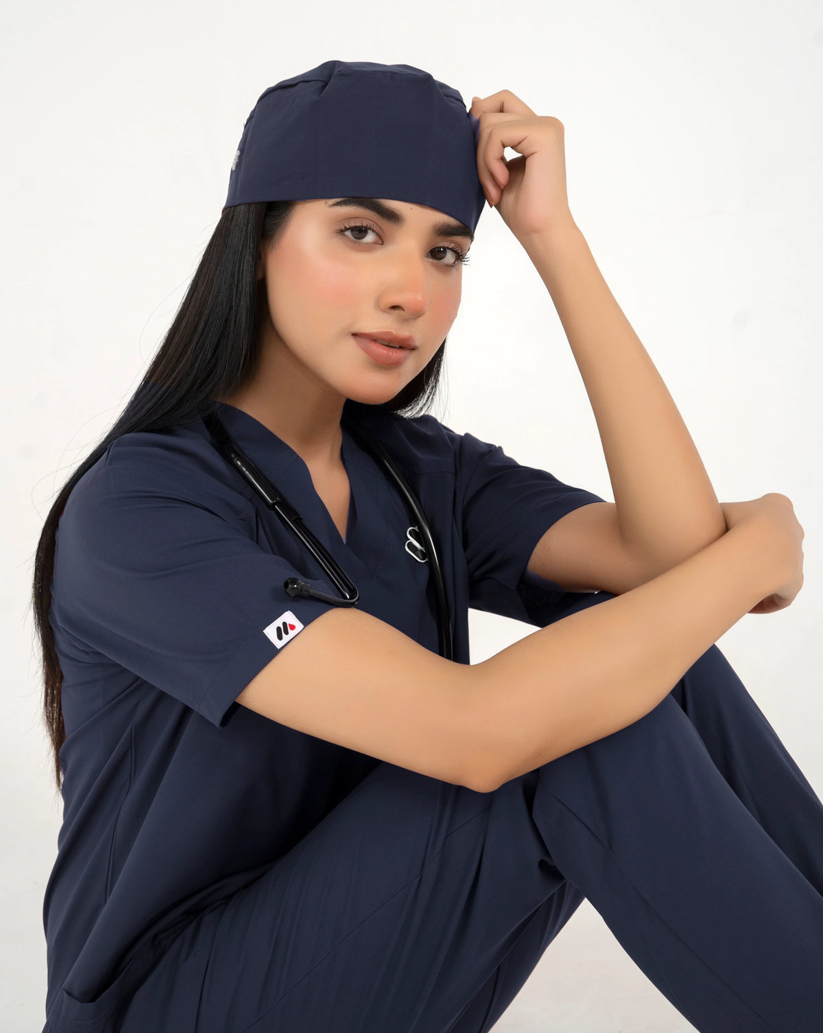 A healthcare professional wearing a matching navy blue medical scrub set with a matching scrub cap and a stethoscope, posing against a white studio background.