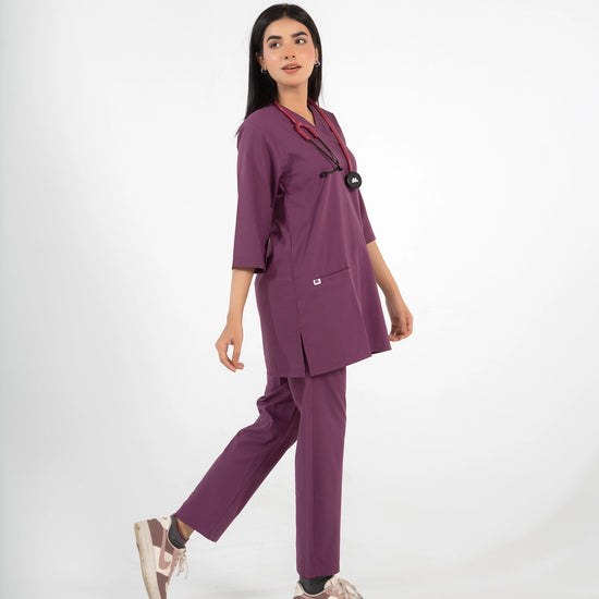 A healthcare professional walking and modeling a purple medical scrub set with a V-neck top, matching pants, a stethoscope, and sneakers against a white background.