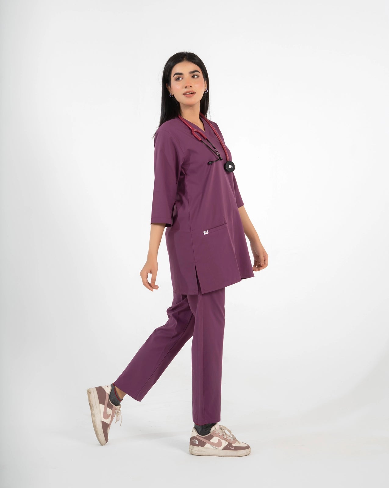 A healthcare professional walking and modeling a purple medical scrub set with a V-neck top, matching pants, a stethoscope, and sneakers against a white background.