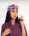 A healthcare professional wearing a purple V-neck medical scrub top, an enamel lapel pin, a stethoscope, and a purple medical scrub cap featuring a playful tooth pattern against a white background.
