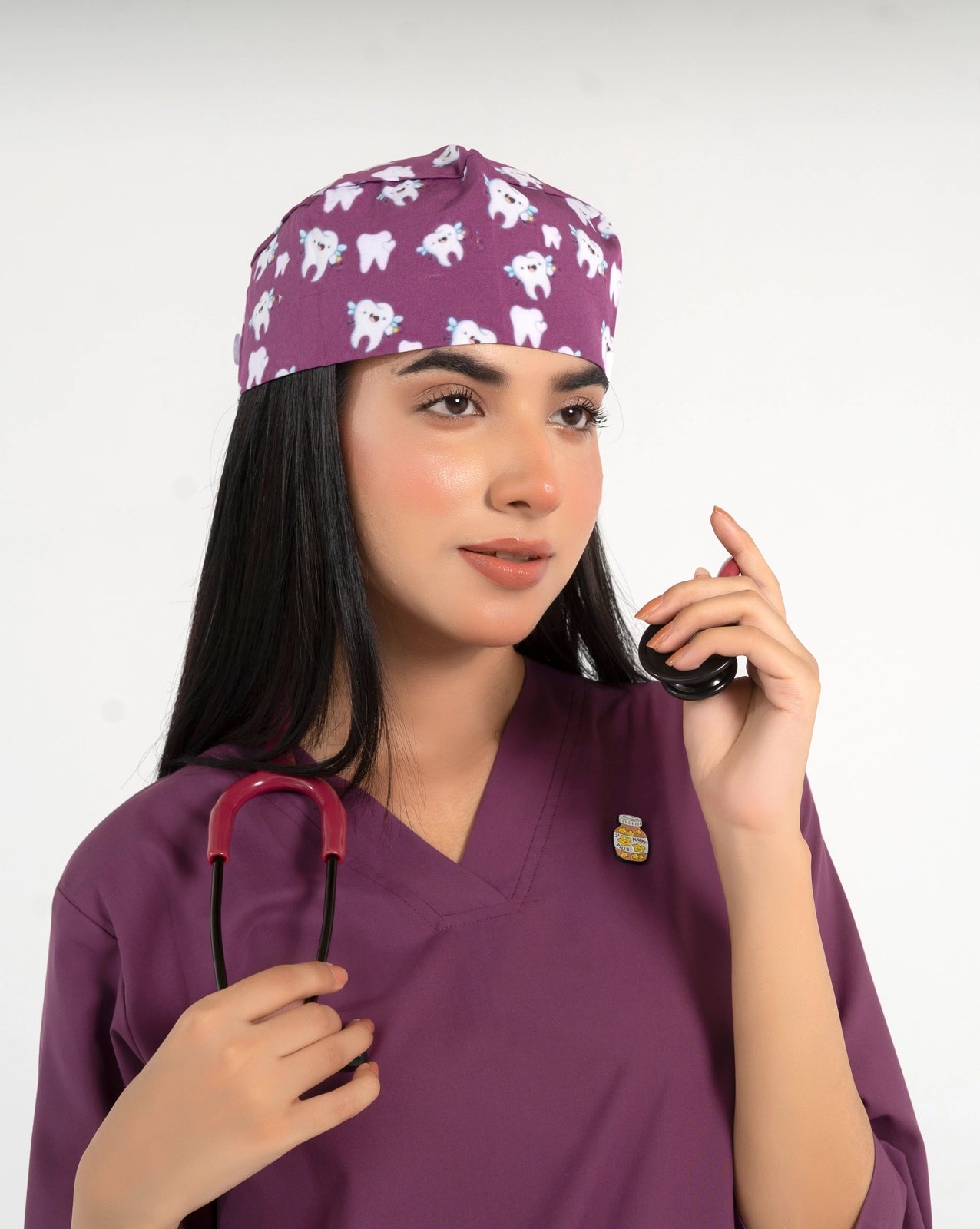 A healthcare professional wearing a purple V-neck medical scrub top, an enamel lapel pin, a stethoscope, and a purple medical scrub cap featuring a playful tooth pattern against a white background.