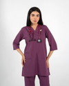A healthcare professional wearing a purple V-neck medical scrub top with a lapel pin and a stethoscope around her neck against a white background.