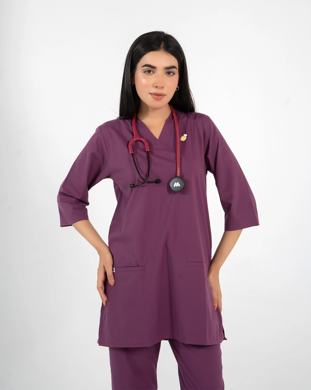 A healthcare professional wearing a purple V-neck medical scrub top with a lapel pin and a stethoscope around her neck against a white background.